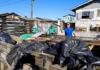 Criciúma promove mutirão de limpeza no bairro Progresso Voluntários e servidores municipais em Criciúma carregando a caçamba de um caminhão de madeira com sacos de lixo e detritos durante o Mutirão de Limpeza do programa "Criciúma, quem ama cuida" no bairro Progresso. Um homem com camisa de time azul levanta uma tábua, enquanto outros dois com uniformes verdes trabalham ao fundo. A cena é uma rua residencial sob céu limpo.