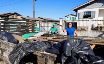 Criciúma promove mutirão de limpeza no bairro Progresso Voluntários e servidores municipais em Criciúma carregando a caçamba de um caminhão de madeira com sacos de lixo e detritos durante o Mutirão de Limpeza do programa "Criciúma, quem ama cuida" no bairro Progresso. Um homem com camisa de time azul levanta uma tábua, enquanto outros dois com uniformes verdes trabalham ao fundo. A cena é uma rua residencial sob céu limpo.