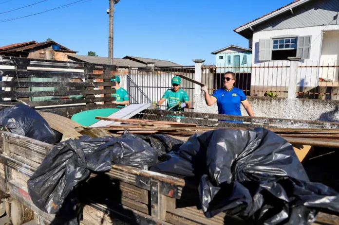 Criciúma promove mutirão de limpeza no bairro Progresso Voluntários e servidores municipais em Criciúma carregando a caçamba de um caminhão de madeira com sacos de lixo e detritos durante o Mutirão de Limpeza do programa "Criciúma, quem ama cuida" no bairro Progresso. Um homem com camisa de time azul levanta uma tábua, enquanto outros dois com uniformes verdes trabalham ao fundo. A cena é uma rua residencial sob céu limpo.