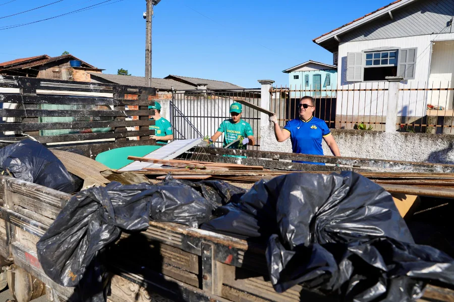 Criciúma promove mutirão de limpeza no bairro Progresso Voluntários e servidores municipais em Criciúma carregando a caçamba de um caminhão de madeira com sacos de lixo e detritos durante o Mutirão de Limpeza do programa