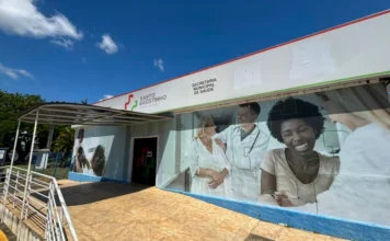 Criciúma terá novo Centro de Diagnóstico de Imagem Uma fotografia da fachada do Complexo de Saúde Santo Agostinho, localizado no Distrito de Rio Maina, Criciúma. O prédio, que abriga a Secretaria Municipal de Saúde conforme o letreiro superior ao lado de um logotipo médico estilizado, exibe uma rampa de acessibilidade com corrimão de metal levando à entrada sob um toldo. A parede externa principal é adornada por um grande e vibrante mural fotográfico mostrando uma idosa sorridente sendo atendida por um jovem médico, ao lado de uma jovem mulher negra também sorrindo largamente. A cena se passa em um dia ensolarado com céu azul e nuvens brancas, com árvores frondosas à esquerda. Este é o local onde está sendo implantado o novo Centro de Diagnóstico de Imagem de Criciúma.