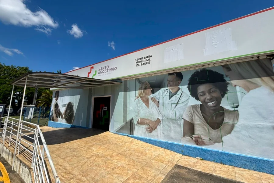Uma fotografia da fachada do Complexo de Saúde Santo Agostinho, localizado no Distrito de Rio Maina, Criciúma. O prédio, que abriga a Secretaria Municipal de Saúde conforme o letreiro superior ao lado de um logotipo médico estilizado, exibe uma rampa de acessibilidade com corrimão de metal levando à entrada sob um toldo. A parede externa principal é adornada por um grande e vibrante mural fotográfico mostrando uma idosa sorridente sendo atendida por um jovem médico, ao lado de uma jovem mulher negra também sorrindo largamente. A cena se passa em um dia ensolarado com céu azul e nuvens brancas, com árvores frondosas à esquerda. Este é o local onde está sendo implantado o novo Centro de Diagnóstico de Imagem de Criciúma.