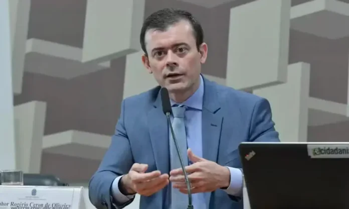 Dario Durigan define nova cúpula do Ministério da Fazenda Rogério Ceron, novo secretário-executivo do Ministério da Fazenda, de terno azul, gesticulando enquanto fala ao microfone em uma sessão oficial em Brasília.