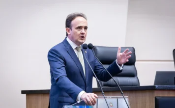 Deputados criticam plano de obras para a BR-101 Norte Um plano médio do deputado Carlos Humberto (PL) discursando na tribuna da Assembleia Legislativa de Santa Catarina (Alesc), em Florianópolis. Ele está de pé atrás de um púlpito de madeira e granito escuro, que tem um brasão de metal na frente. O deputado veste um terno azul-marinho, camisa branca e gravata clara estampada em tons de bege. Ele está falando, gesticulando ativamente com a mão direita erguida, enquanto a mão esquerda está apoiada sobre o púlpito. Há dois microfones em hastes flexíveis à sua frente. O fundo desfocado mostra cadeiras de couro pretas e a borda de um laptop sobre uma bancada. O deputado fala sobre a falta de investimentos na repactuação da BR-101.