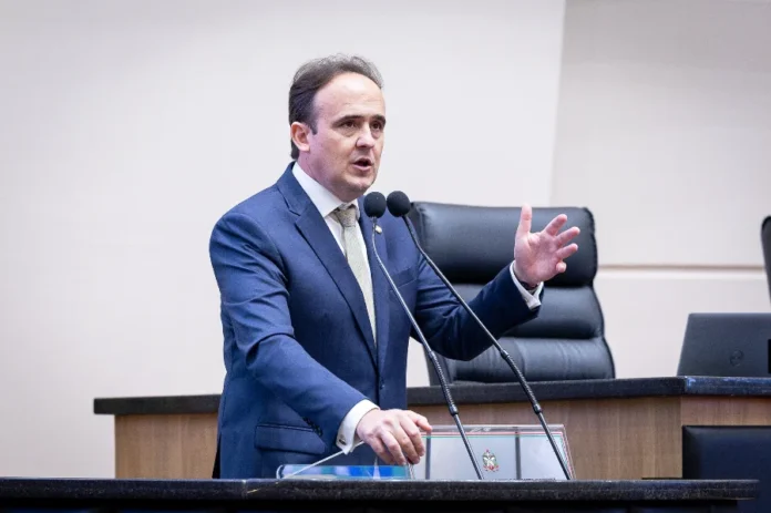 Deputados criticam plano de obras para a BR-101 Norte Um plano médio do deputado Carlos Humberto (PL) discursando na tribuna da Assembleia Legislativa de Santa Catarina (Alesc), em Florianópolis. Ele está de pé atrás de um púlpito de madeira e granito escuro, que tem um brasão de metal na frente. O deputado veste um terno azul-marinho, camisa branca e gravata clara estampada em tons de bege. Ele está falando, gesticulando ativamente com a mão direita erguida, enquanto a mão esquerda está apoiada sobre o púlpito. Há dois microfones em hastes flexíveis à sua frente. O fundo desfocado mostra cadeiras de couro pretas e a borda de um laptop sobre uma bancada. O deputado fala sobre a falta de investimentos na repactuação da BR-101.