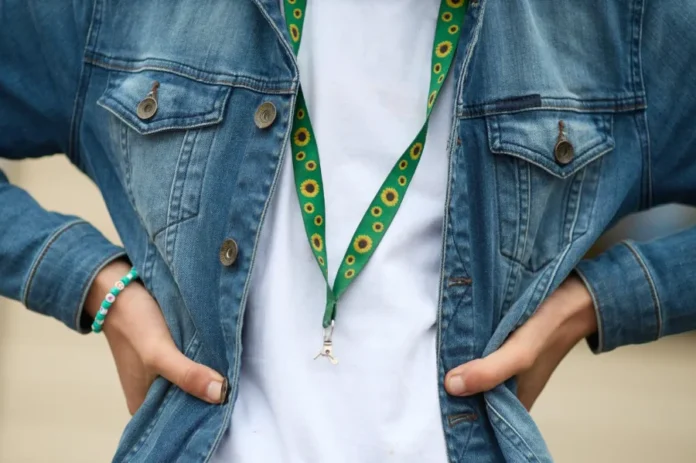 Deputados de SC aprovam uso oficial do Cordão de Girassol Um close-up focado no tronco de uma pessoa que veste uma jaqueta jeans azul de lavagem média, aberta, sobre uma camiseta branca lisa. A pessoa está com as mãos nos quadris, com os dedos enfiados nos bolsos da jaqueta. No pescoço, ela usa um cordão (lanyard) de tecido verde vibrante com um padrão repetitivo de girassóis amarelos com centros escuros, que é o símbolo reconhecido internacionalmente para deficiências ocultas. O cordão tem um pequeno clipe de metal prateado e um gancho na extremidade, mas não exibe nenhum crachá ou ID específico. No pulso esquerdo, a pessoa usa uma pulseira de miçangas turquesa e branca. O fundo é uma parede de estuque em tom creme/amarelo pálido. Esta imagem ilustra o Cordão de Girassol, agora oficializado como ferramenta de identificação em Santa Catarina.