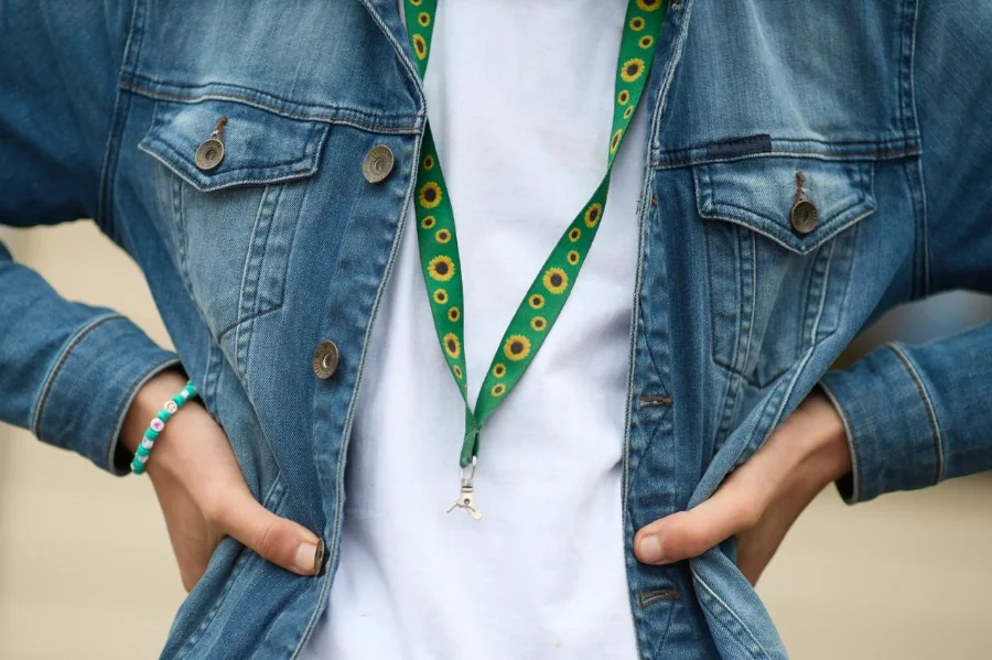 Um close-up focado no tronco de uma pessoa que veste uma jaqueta jeans azul de lavagem média, aberta, sobre uma camiseta branca lisa. A pessoa está com as mãos nos quadris, com os dedos enfiados nos bolsos da jaqueta. No pescoço, ela usa um cordão (lanyard) de tecido verde vibrante com um padrão repetitivo de girassóis amarelos com centros escuros, que é o símbolo reconhecido internacionalmente para deficiências ocultas. O cordão tem um pequeno clipe de metal prateado e um gancho na extremidade, mas não exibe nenhum crachá ou ID específico. No pulso esquerdo, a pessoa usa uma pulseira de miçangas turquesa e branca. O fundo é uma parede de estuque em tom creme/amarelo pálido. Esta imagem ilustra o Cordão de Girassol, agora oficializado como ferramenta de identificação em Santa Catarina.