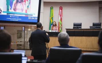 Deputados debatem combate à violência contra a mulher Uma fotografia tirada de dentro do plenário da Assembleia Legislativa de Santa Catarina (Alesc), com um ponto de vista de trás de outras pessoas desfocadas na plateia ou área de mídia. No centro, a Deputada Ana Campagnolo (com cabelos ruivos e vestindo uma blusa xadrez vermelha e branca) fala ao microfone do púlpito de madeira. Atrás dela, estão dispostas as bandeiras do Brasil e de Santa Catarina. À esquerda, uma grande tela de monitor de vídeo exibe um close-up da oradora e o texto legível identificando-a explicitamente: "Deputada Ana Campagnolo", o tema "Mês da Mulher", o evento "15ª SESSÃO PLENÁRIA ORDINÁRIA", a data "5 DE MARÇO DE 2026" e o tempo de fala de "00:04:08". O ambiente é formal, com móveis de madeira e painéis acústicos.
