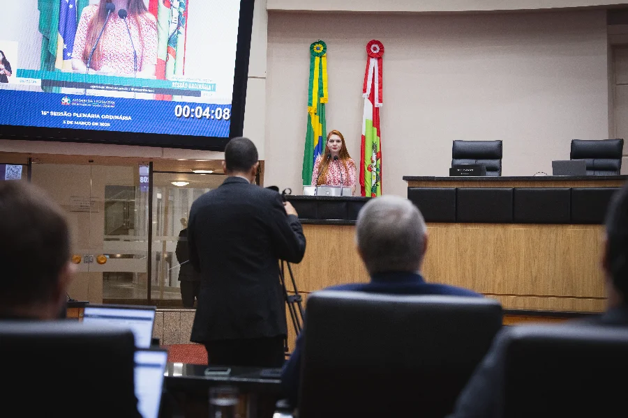 Uma fotografia tirada de dentro do plenário da Assembleia Legislativa de Santa Catarina (Alesc), com um ponto de vista de trás de outras pessoas desfocadas na plateia ou área de mídia. No centro, a Deputada Ana Campagnolo (com cabelos ruivos e vestindo uma blusa xadrez vermelha e branca) fala ao microfone do púlpito de madeira. Atrás dela, estão dispostas as bandeiras do Brasil e de Santa Catarina. À esquerda, uma grande tela de monitor de vídeo exibe um close-up da oradora e o texto legível identificando-a explicitamente: 