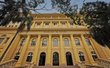 Diálogos Setoriais debatem desempenho da indústria no RS Fotografia de ângulo baixo da imponente fachada neoclássica amarela do edifício da Secretaria da Fazenda. Colunas coríntias grandiosas apoiam a varanda superior, onde a inscrição "SECRETARIA DA FAZENDA" está gravada. A entrada principal tem três grandes portas em arco de ferro forjado no topo de uma escadaria de pedra. Bandeiras do Brasil e do Rio Grande do Sul estão hasteadas. A foto é emoldurada por frondosos galhos de árvores contra um céu azul limpo.