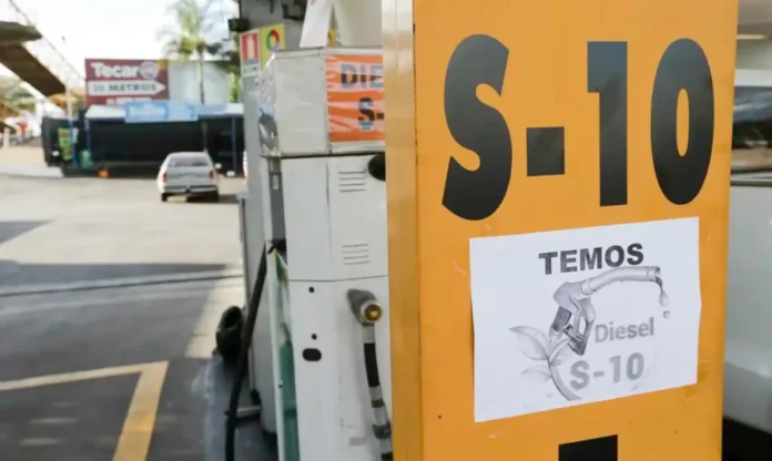 Diesel em falta ameaça colheita de arroz e soja no RS Totem vertical amarelo de um posto de gasolina em primeiro plano, com o texto grande em preto "S-10" no topo. Abaixo, um cartaz de papel impresso colado com fita adesiva que diz: "TEMOS" acima de uma ilustração de um bico de abastecimento de combustível com folhas verdes estilizadas, com "Diesel" e "S-10" abaixo. O fundo desfocado mostra a bomba de combustível branca, uma mangueira e bico de abastecimento, um carro sedan estacionado e um letreiro de "Tecar 20 METROS".