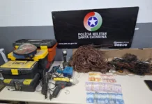 Dois homens são detidos com fios e balança em Criciúma Foto de evidências policiais apreendidas em Criciúma. Exposição de um grande monte de fios de cobre desencapados ao lado de caixas de ferramentas empilhadas, furadeiras e outras ferramentas elétricas. Há também diversas cédulas de dinheiro em Real e um banner oficial da Polícia Militar de Santa Catarina (PMSC) ao fundo.