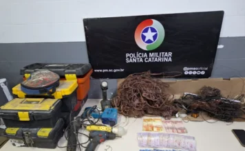 Dois homens são detidos com fios e balança em Criciúma Foto de evidências policiais apreendidas em Criciúma. Exposição de um grande monte de fios de cobre desencapados ao lado de caixas de ferramentas empilhadas, furadeiras e outras ferramentas elétricas. Há também diversas cédulas de dinheiro em Real e um banner oficial da Polícia Militar de Santa Catarina (PMSC) ao fundo.