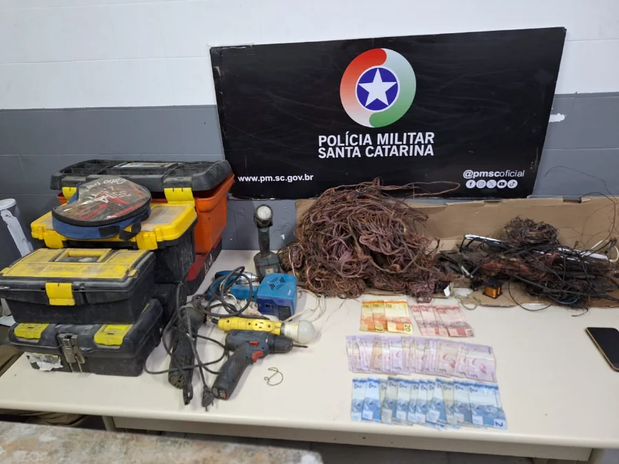 Dois homens são detidos com fios e balança em Criciúma Foto de evidências policiais apreendidas em Criciúma. Exposição de um grande monte de fios de cobre desencapados ao lado de caixas de ferramentas empilhadas, furadeiras e outras ferramentas elétricas. Há também diversas cédulas de dinheiro em Real e um banner oficial da Polícia Militar de Santa Catarina (PMSC) ao fundo.