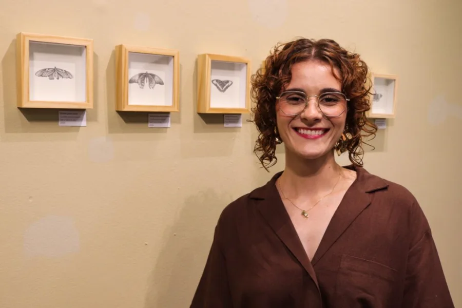 Elisa Corrêa abre mostra Ensaio sobre Coleções na Unesc A artista visual Elisa Corrêa sorri em frente a quatro quadros com molduras claras contendo desenhos detalhados de mariposas em grafite, durante a abertura da exposição