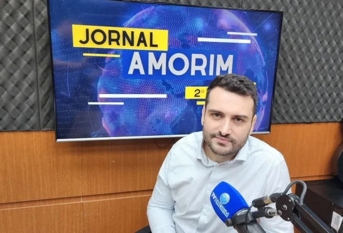 Entenda o impacto da reforma nos benefícios do INSS O advogado Rafael Furlanetto de Nes, um homem de pele clara, cabelos e barba escuros e curtos, veste uma camisa social de cor clara. Ele está sentado em um estúdio de rádio profissional, posicionado atrás de um microfone azul com a marca "TV Amorim". Ao fundo, uma tela de televisão exibe o logotipo do "Jornal Amorim" sobre um gráfico global.