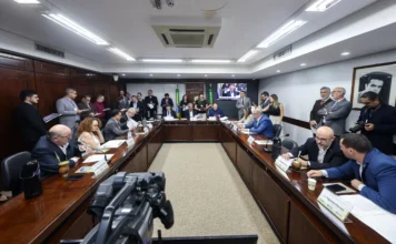 EstruTUR-RS: PEC do Turismo avança na Assembleia Gaúcha Fotografia em ângulo amplo de uma reunião formal da Comissão de Constituição e Justiça (CCJ) do Brasil, no Congresso Nacional, em Brasília. Parlamentares e autoridades estão sentados em uma mesa de conferência em U, com o brasão da CCJ e telas de transmissão ao vivo ao fundo. Uma câmera de vídeo profissional de broadcast está em primeiro plano, registrando a sessão legislativa e a cobertura de imprensa.