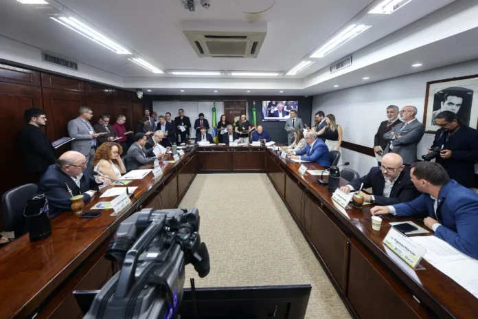 EstruTUR-RS PEC do Turismo avança na Assembleia Gaúcha Fotografia em ângulo amplo de uma reunião formal da Comissão de Constituição e Justiça (CCJ) do Brasil, no Congresso Nacional, em Brasília. Parlamentares e autoridades estão sentados em uma mesa de conferência em U, com o brasão da CCJ e telas de transmissão ao vivo ao fundo. Uma câmera de vídeo profissional de broadcast está em primeiro plano, registrando a sessão legislativa e a cobertura de imprensa.