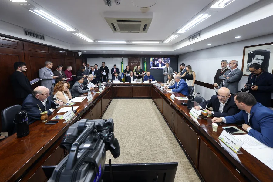 Fotografia em ângulo amplo de uma reunião formal da Comissão de Constituição e Justiça (CCJ) do Brasil, no Congresso Nacional, em Brasília. Parlamentares e autoridades estão sentados em uma mesa de conferência em U, com o brasão da CCJ e telas de transmissão ao vivo ao fundo. Uma câmera de vídeo profissional de broadcast está em primeiro plano, registrando a sessão legislativa e a cobertura de imprensa.