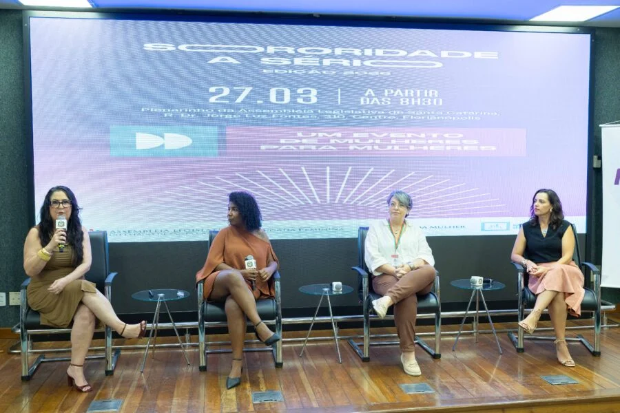 Evento na Alesc incentiva o empreendedorismo feminino Painel de debates com quatro mulheres sentadas no palco do Plenarinho da Alesc durante o evento Sororidade a Sério Edição 2023. O telão de fundo exibe o título do evento, a data 27.03 e a frase 'Um evento de mulheres para mulheres'. Mulheres brancas e negras compõem a mesa, com uma delas falando ao microfone. Foco em empreendedorismo feminino e liderança.