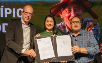 Famurs e Assembleia do RS assinam pacto pelo municipalismo Foto de palco: o deputado Sergio Peres, presidente da Assembleia Legislativa do RS (esquerda), a prefeita Adriane Perin de Oliveira, presidente da Famurs (centro), e o deputado Joel Wilhem (direita) seguram o Termo de Cooperação Técnica assinado em Torres. Ao fundo, uma tela de LED exibe a imagem de um agricultor e a palavra parcial "MUNICÍPIO", contextualizando a união das pautas legislativas e municipais.