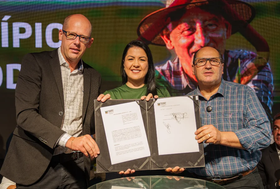 Foto de palco: o deputado Sergio Peres, presidente da Assembleia Legislativa do RS (esquerda), a prefeita Adriane Perin de Oliveira, presidente da Famurs (centro), e o deputado Joel Wilhem (direita) seguram o Termo de Cooperação Técnica assinado em Torres. Ao fundo, uma tela de LED exibe a imagem de um agricultor e a palavra parcial 