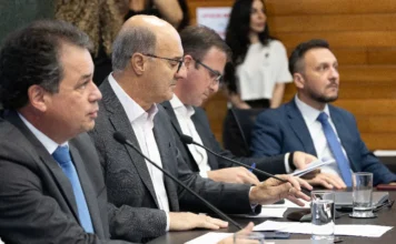 Plano para desassorear Rio Urussanga avança na Alesc Uma foto de arquivo horizontal em plano médio captura quatro homens formalmente vestidos com ternos e gravatas, sentados em uma longa mesa de conferência escura na sala de comissões da Alesc. O homem no centro, calvo e de óculos, segura documentos e fala ao microfone de mesa, cercado pelos outros participantes focados em seus papéis. A profundidade de campo é rasa, mostrando uma mulher desfocada ao fundo. A imagem ilustra a instalação da Frente Parlamentar em Defesa da Limpeza e Desassoreamento do Rio Urussanga.