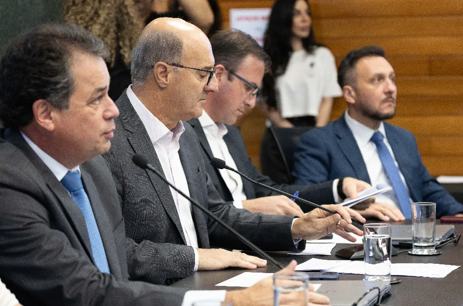 Uma foto de arquivo horizontal em plano médio captura quatro homens formalmente vestidos com ternos e gravatas, sentados em uma longa mesa de conferência escura na sala de comissões da Alesc. O homem no centro, calvo e de óculos, segura documentos e fala ao microfone de mesa, cercado pelos outros participantes focados em seus papéis. A profundidade de campo é rasa, mostrando uma mulher desfocada ao fundo. A imagem ilustra a instalação da Frente Parlamentar em Defesa da Limpeza e Desassoreamento do Rio Urussanga.