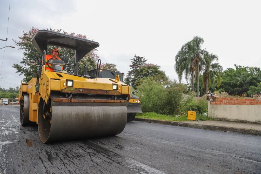 Gabinete Itinerante de Criciúma leva melhorias à Cidade Mineira Rolo compressor amarelo da DTT realiza pavimentação asfáltica em rua da região da Cidade Mineira, Criciúma, durante ação do Gabinete Itinerante. Máquina compacta asfalto novo sobre paralelepípedos com operador visível.