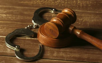 Homem é condenado a 38 anos por feminicídio em Turvo A imagem mostra um close-up focado de um martelo de juiz de madeira com seu bloco de ressonância, posicionado ao lado de um par de algemas de metal abertas sobre uma superfície de madeira texturizada. Esta cena simboliza o resultado de um julgamento, representando a conclusão de um processo judicial e a consequente punição, como a condenação de um réu por feminicídio a uma pena de reclusão de 38 anos em regime fechado.