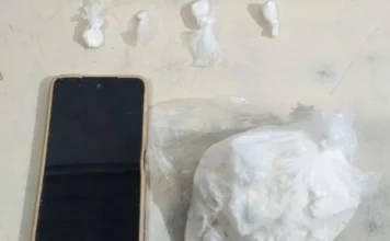 Homem é preso com 250g de cocaína no Arroio do Silva Fotografia em plano superior sobre uma superfície clara exibe os itens apreendidos: à esquerda, um smartphone de tela preta e capa bege; acima, quatro pequenas porções de substância branca embaladas individualmente em plástico; à direita, um saco plástico transparente contendo uma pedra grande de substância branca análoga à cocaína.