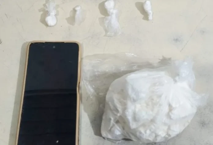 Homem é preso com 250g de cocaína no Arroio do Silva Fotografia em plano superior sobre uma superfície clara exibe os itens apreendidos: à esquerda, um smartphone de tela preta e capa bege; acima, quatro pequenas porções de substância branca embaladas individualmente em plástico; à direita, um saco plástico transparente contendo uma pedra grande de substância branca análoga à cocaína.