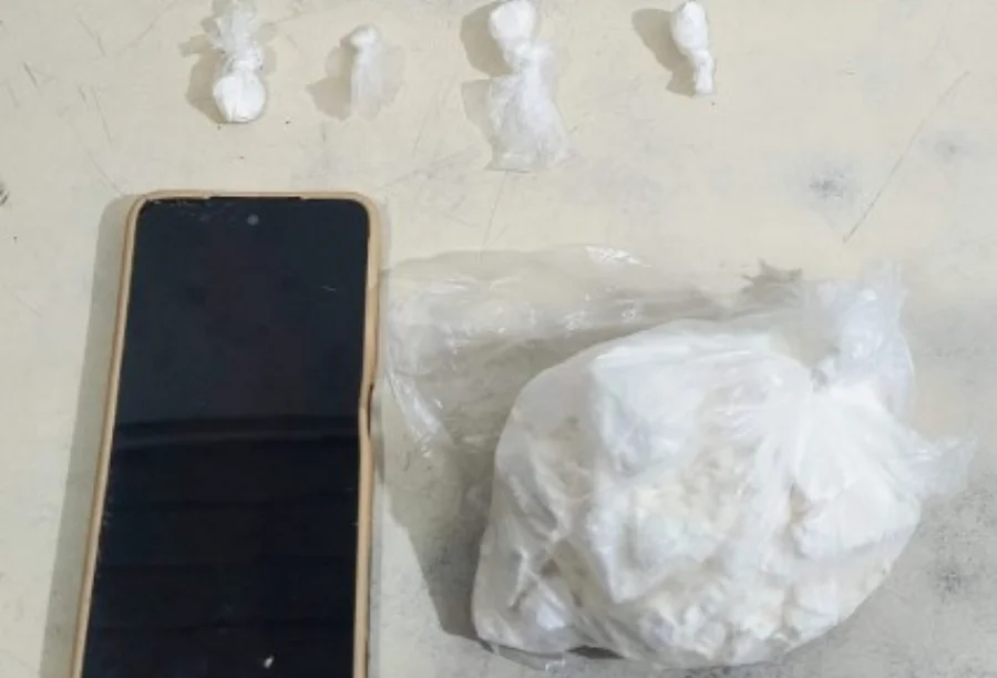 Fotografia em plano superior sobre uma superfície clara exibe os itens apreendidos: à esquerda, um smartphone de tela preta e capa bege; acima, quatro pequenas porções de substância branca embaladas individualmente em plástico; à direita, um saco plástico transparente contendo uma pedra grande de substância branca análoga à cocaína.