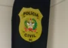 Imagem de close-up de um emblema de tecido oval, de cor amarela, fixado a um pano preto pendurado no canto de uma parede branca. O emblema traz o texto 'POLÍCIA' na parte superior e 'CIVIL' na parte inferior, com 'SC' abaixo, tudo em letras pretas maiúsculas. No centro do emblema amarelo, há um brasão heráldico detalhado, composto por elementos dourados e vermelhos sobre um fundo branco.