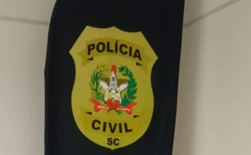 Homem é preso por violência doméstica em Araranguá Imagem de close-up de um emblema de tecido oval, de cor amarela, fixado a um pano preto pendurado no canto de uma parede branca. O emblema traz o texto 'POLÍCIA' na parte superior e 'CIVIL' na parte inferior, com 'SC' abaixo, tudo em letras pretas maiúsculas. No centro do emblema amarelo, há um brasão heráldico detalhado, composto por elementos dourados e vermelhos sobre um fundo branco.