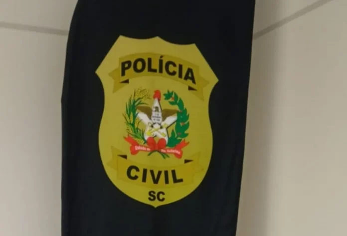 Homem é preso por violência doméstica em Araranguá Imagem de close-up de um emblema de tecido oval, de cor amarela, fixado a um pano preto pendurado no canto de uma parede branca. O emblema traz o texto 'POLÍCIA' na parte superior e 'CIVIL' na parte inferior, com 'SC' abaixo, tudo em letras pretas maiúsculas. No centro do emblema amarelo, há um brasão heráldico detalhado, composto por elementos dourados e vermelhos sobre um fundo branco.