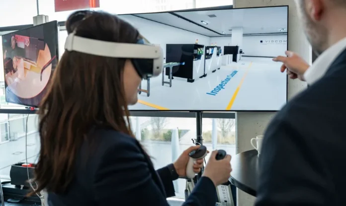 IA na indústria 95% dos projetos ainda não geram lucro Uma mulher de cabelos longos, vista de costas, usa um headset de realidade virtual branco e segura controles nas duas mãos, interagindo com uma grande tela de monitor que exibe um ambiente de fábrica virtual com máquinas modulares da VISCOM e o texto "Inspection Cellule" no chão. À sua direita, um homem de terno está de pé e aponta com o dedo para a tela. Um monitor secundário menor à esquerda mostra uma visão externa da mesma demonstração de VR. A cena ocorre em um pavilhão de exposição.