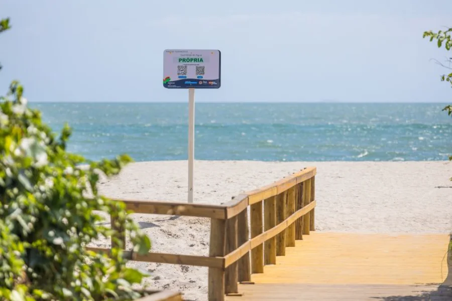 Uma passarela de madeira em primeiro plano conduz o olhar para uma faixa de areia clara e o mar azul ao fundo, sob céu aberto. No centro da imagem, em destaque, há uma placa branca de sinalização instalada pelo IMA. No topo da placa, a palavra 