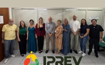IPREV recebe R$ 67,4 milhões em compensação previdenciária Uma foto de grupo oficial com nove funcionários do IPREV (Instituto de Previdência de Santa Catarina) em pé, lado a lado, em um ambiente de escritório com divisórias de vidro jateado e piso de azulejo branco. Os nove integrantes (cinco mulheres e quatro homens) estão sorrindo para a câmera. O homem no centro usa uma camisa cinza, ladeado por mulheres em vestidos longos (azul turquesa, azul marinho e estampado) e uma mulher em blazer vermelho. Nas extremidades, outros membros da equipe usam roupas casuais (camisetas e jeans) e trajes de trabalho. No primeiro plano inferior, está sobreposto o logotipo do IPREV, composto por três formas ovais coloridas (vermelho, verde e amarelo) e a palavra 'IPREV' em letras verdes escuras.