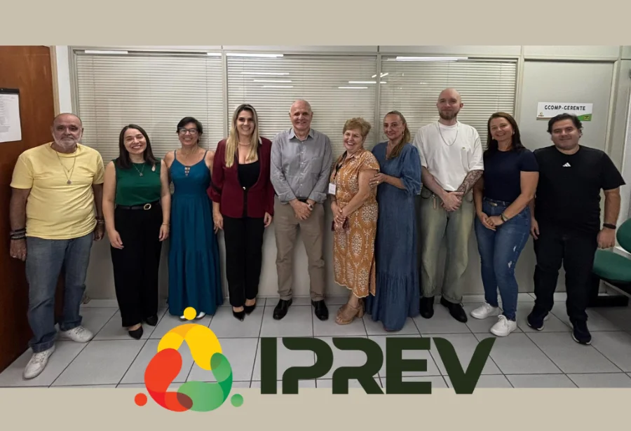 Uma foto de grupo oficial com nove funcionários do IPREV (Instituto de Previdência de Santa Catarina) em pé, lado a lado, em um ambiente de escritório com divisórias de vidro jateado e piso de azulejo branco. Os nove integrantes (cinco mulheres e quatro homens) estão sorrindo para a câmera. O homem no centro usa uma camisa cinza, ladeado por mulheres em vestidos longos (azul turquesa, azul marinho e estampado) e uma mulher em blazer vermelho. Nas extremidades, outros membros da equipe usam roupas casuais (camisetas e jeans) e trajes de trabalho. No primeiro plano inferior, está sobreposto o logotipo do IPREV, composto por três formas ovais coloridas (vermelho, verde e amarelo) e a palavra 'IPREV' em letras verdes escuras.