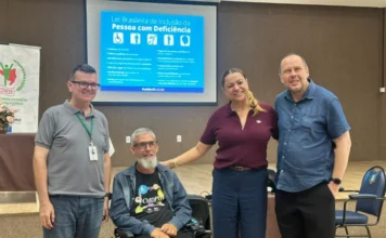 Içara realiza formação sobre direitos e capacitismo Foto de grupo de quatro pessoas sorrindo em um auditório, incluindo um homem usando uma cadeira de rodas no centro com uma camiseta do CMDPD Içara. Atrás deles, uma tela projeta um slide com o título "Lei Brasileira de Inclusão da Pessoa com Deficiência" e logotipos de acessibilidade. Um banner do CONEDE-SC é visível à esquerda.
