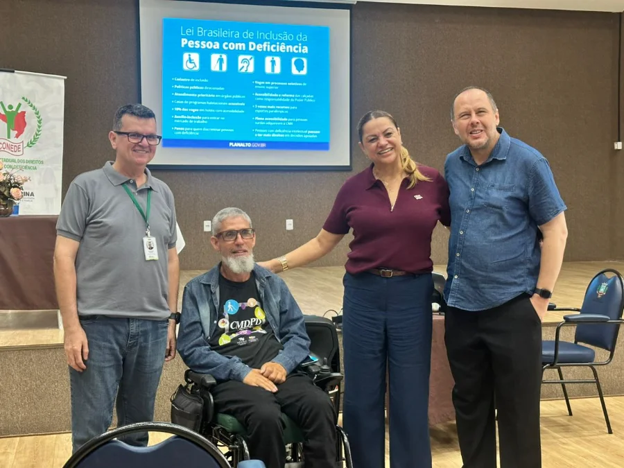 Içara realiza formação sobre direitos e capacitismo Foto de grupo de quatro pessoas sorrindo em um auditório, incluindo um homem usando uma cadeira de rodas no centro com uma camiseta do CMDPD Içara. Atrás deles, uma tela projeta um slide com o título