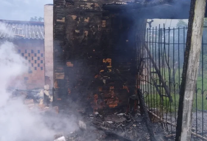 Incêndio destrói casa no bairro Cristo Redentor em Criciúma Uma fotografia horizontal colorida mostra o local de uma residência unifamiliar mista após um incêndio. A parede de tijolos principal está severamente carbonizada, coberta por uma camada espessa de fuligem preta. Fumaça branca densa sobe do chão e se espalha pelo ar no lado esquerdo da imagem, indicando a fase de rescaldo. No chão, escombros carbonizados, destroços e pedaços de madeira e tijolos estão espalhados. À direita, um portão de ferro e uma cerca de arame com postes de concreto delimitam a propriedade, com vegetação densa e uma encosta verde visível ao fundo sob um céu nublado. O telhado da casa adjacente é visível no fundo à esquerda.