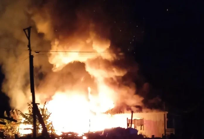 Incêndio destrói residência no Balneário Rincão Fotografia noturna de um incêndio de grandes proporções em uma residência. Chamas intensas de cor laranja e amarela envolvem quase toda a estrutura do imóvel, com uma densa coluna de fumaça escura subindo ao céu. À esquerda, é possível ver a silhueta de um poste de energia e folhagens, enquanto a iluminação do fogo destaca o volume do incêndio no Balneário Rincão.