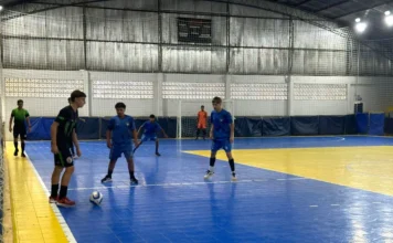 Siderópolis inicia JEMS 2025 com 300 alunos em quadra Jogador de futsal driblando a bola durante uma partida dos Jogos Escolares do Município de Siderópolis (JEMS) 2025. A cena se passa em uma quadra poliesportiva com piso azul e amarelo, mostrando outros jogadores e o árbitro. Foto de ação focada em esportes estudantis.
