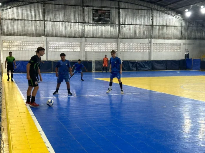 JESSICA ROSSO CREPALDI_PREFEITURA DE SIDERÓPOLIS (1) Jogador de futsal driblando a bola durante uma partida dos Jogos Escolares do Município de Siderópolis (JEMS) 2025. A cena se passa em uma quadra poliesportiva com piso azul e amarelo, mostrando outros jogadores e o árbitro. Foto de ação focada em esportes estudantis.