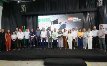 Jorginho Mello entrega 25 ambulâncias e motolâncias em SC Governador Jorginho Mello segura chave simbólica no palco durante cerimônia de entrega de 25 novas ambulâncias e motolâncias para o SAMU em Santa Catarina. Ao fundo, painel exibe a frota de veículos entregues em Içara para reforçar a saúde estadual.