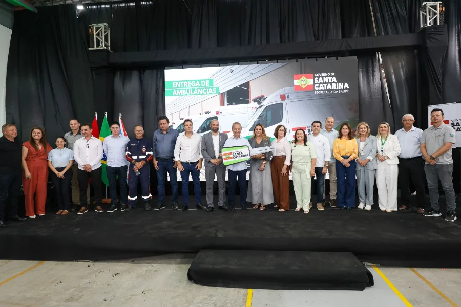 Governador Jorginho Mello segura chave simbólica no palco durante cerimônia de entrega de 25 novas ambulâncias e motolâncias para o SAMU em Santa Catarina. Ao fundo, painel exibe a frota de veículos entregues em Içara para reforçar a saúde estadual.