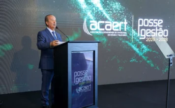 Jorginho Mello prestigia posse da nova diretoria da Acaert O governador de Santa Catarina, Jorginho Mello, aparece de perfil, vestindo terno azul, falando ao microfone em um púlpito preto. Ao fundo, um grande painel digital exibe a logomarca da Acaert (Associação Catarinense de Emissoras de Rádio e Televisão)