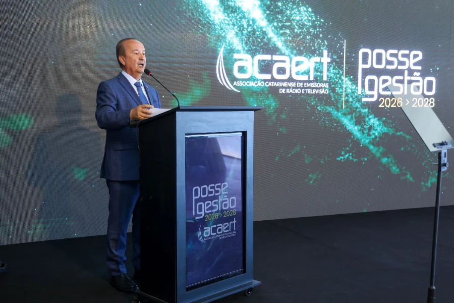 O governador de Santa Catarina, Jorginho Mello, aparece de perfil, vestindo terno azul, falando ao microfone em um púlpito preto. Ao fundo, um grande painel digital exibe a logomarca da Acaert (Associação Catarinense de Emissoras de Rádio e Televisão)